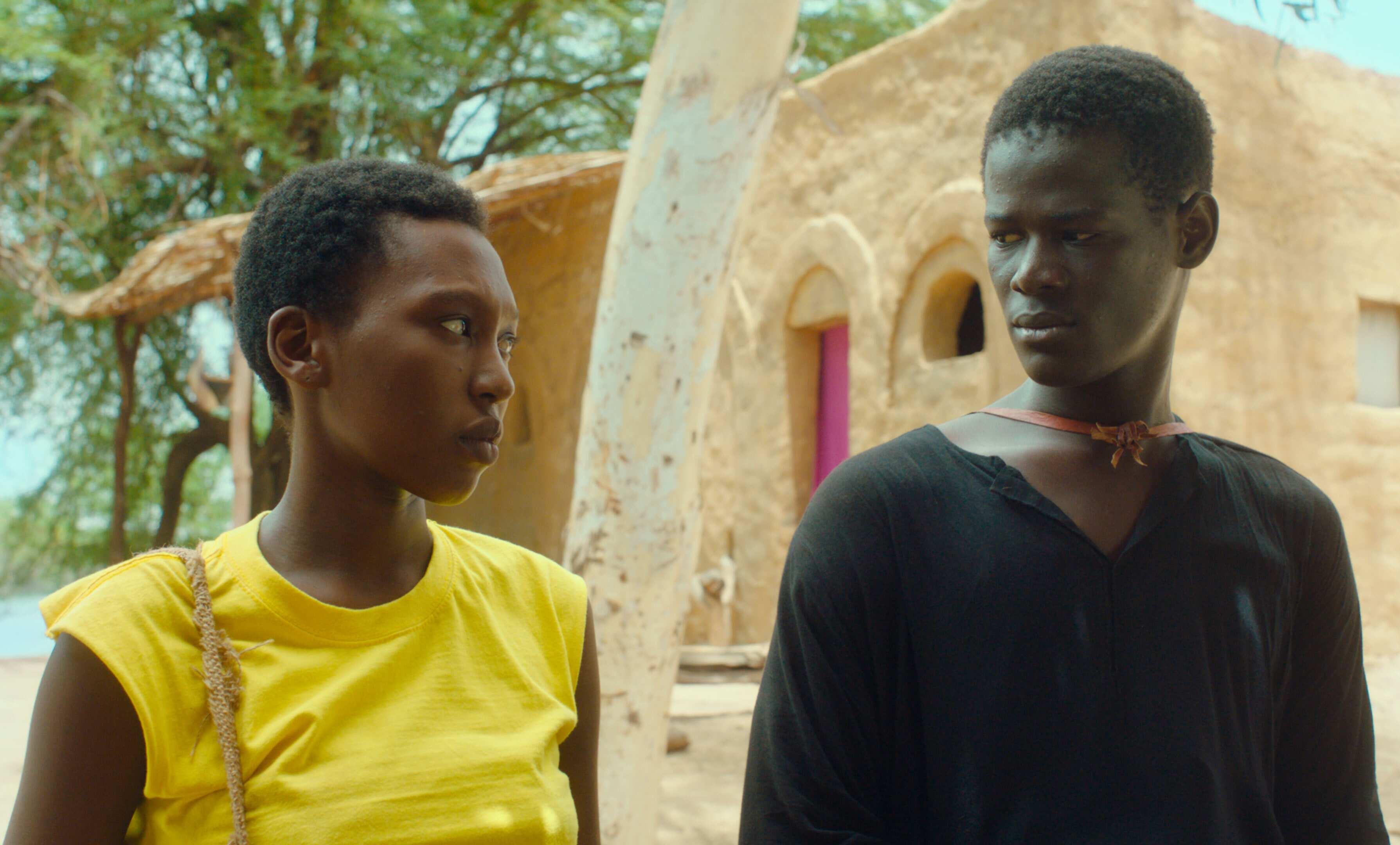 Two people are standing outdoors. One person is wearing a yellow sleeveless top and has a bag strap over one shoulder, while the other is wearing a black long-sleeved shirt and has a necklace around their neck. In the background, there's a rustic building