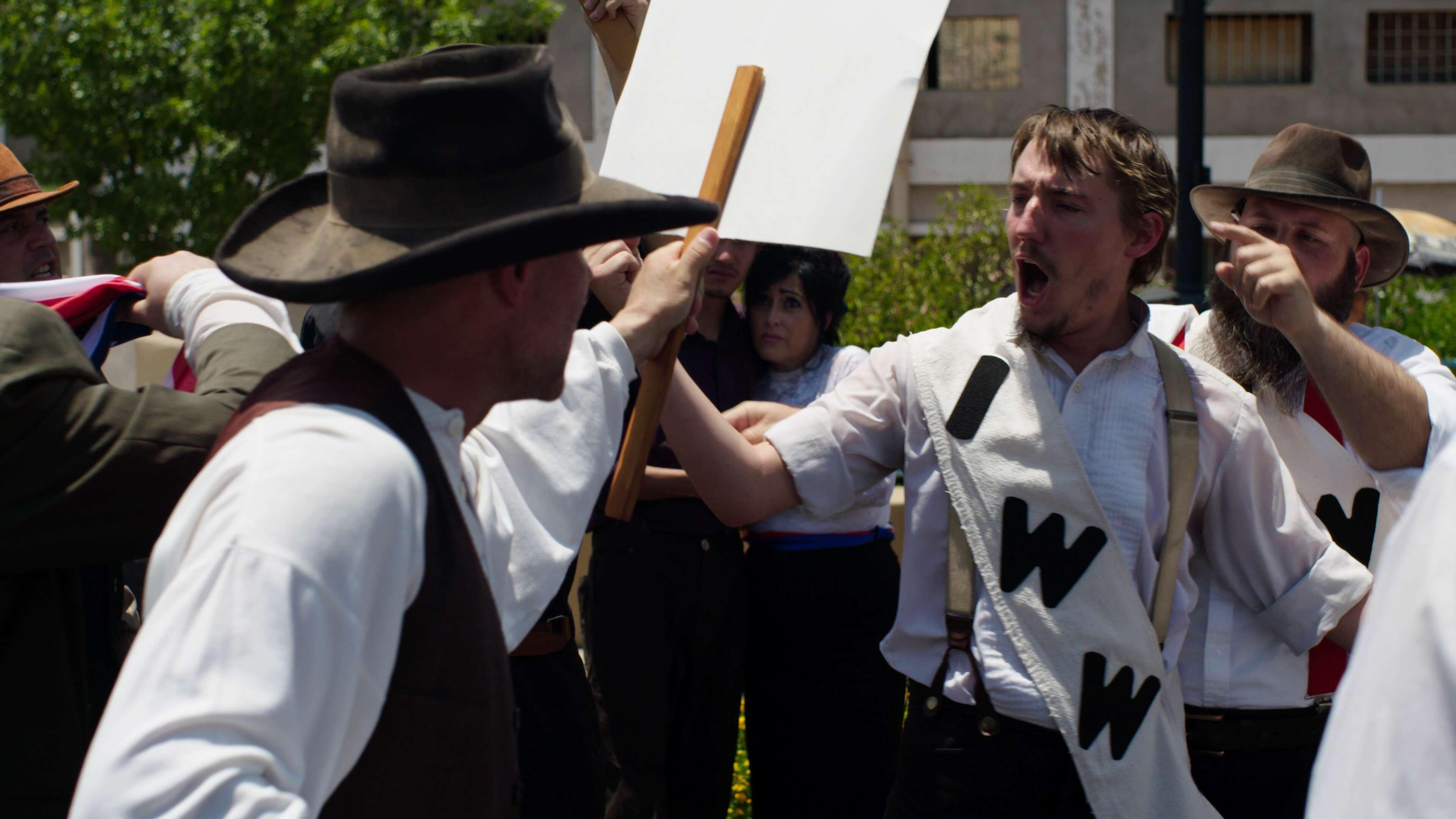 Men in 1917 period clothing gathered outside holding signs and shouting as part of a workers strike.