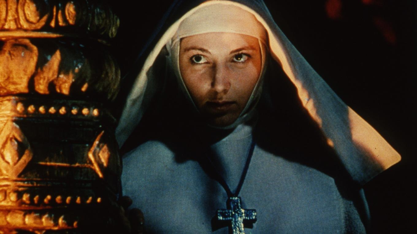 A nun wearing a habit and a cross with her face illuminated by a fire.