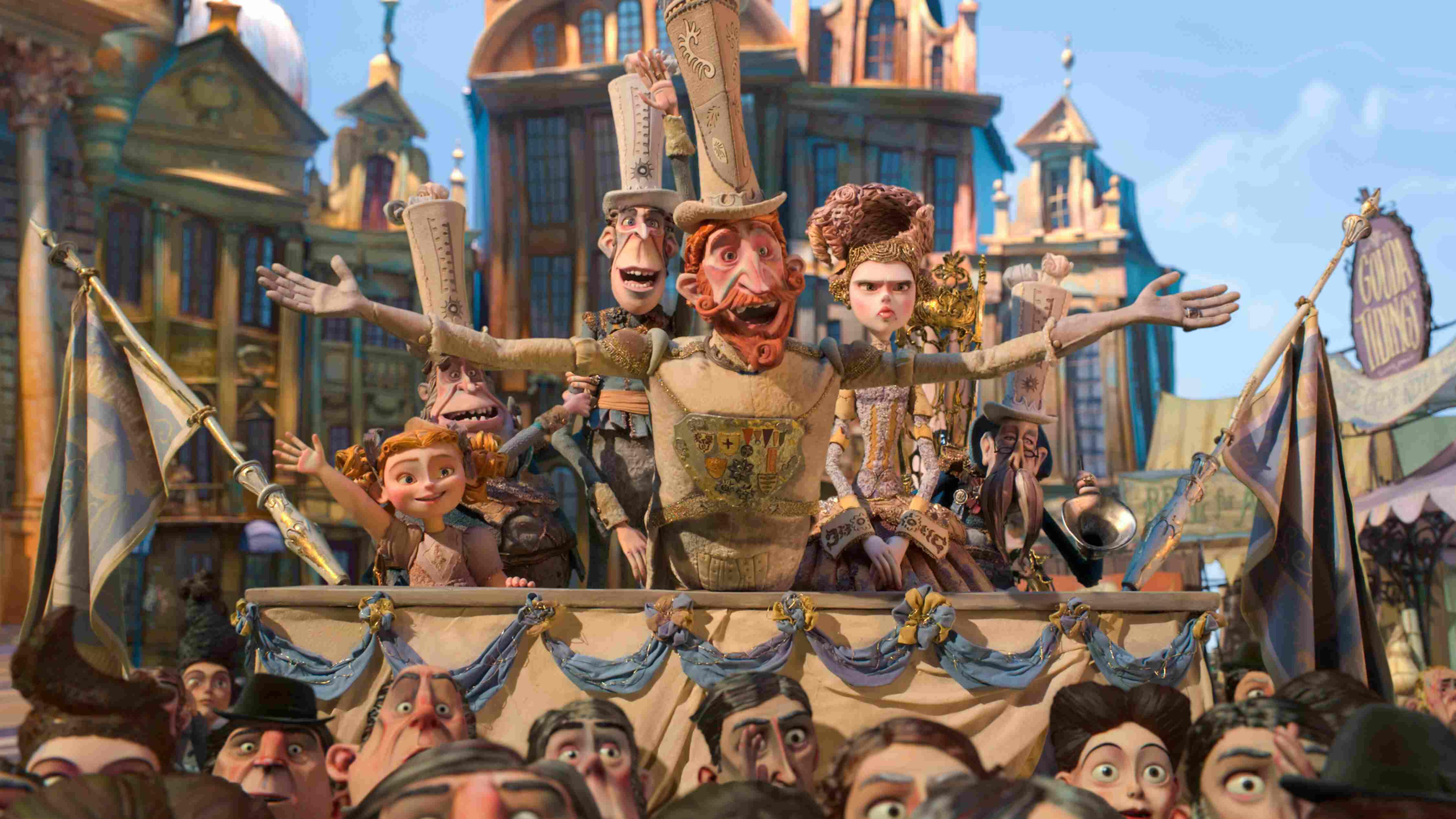 A stop-motion animated scene with a tall figure in a decorated outfit and tall hat, arms outstretched, addressing a crowd in a festive town square, surrounded by characters in elaborate costumes.