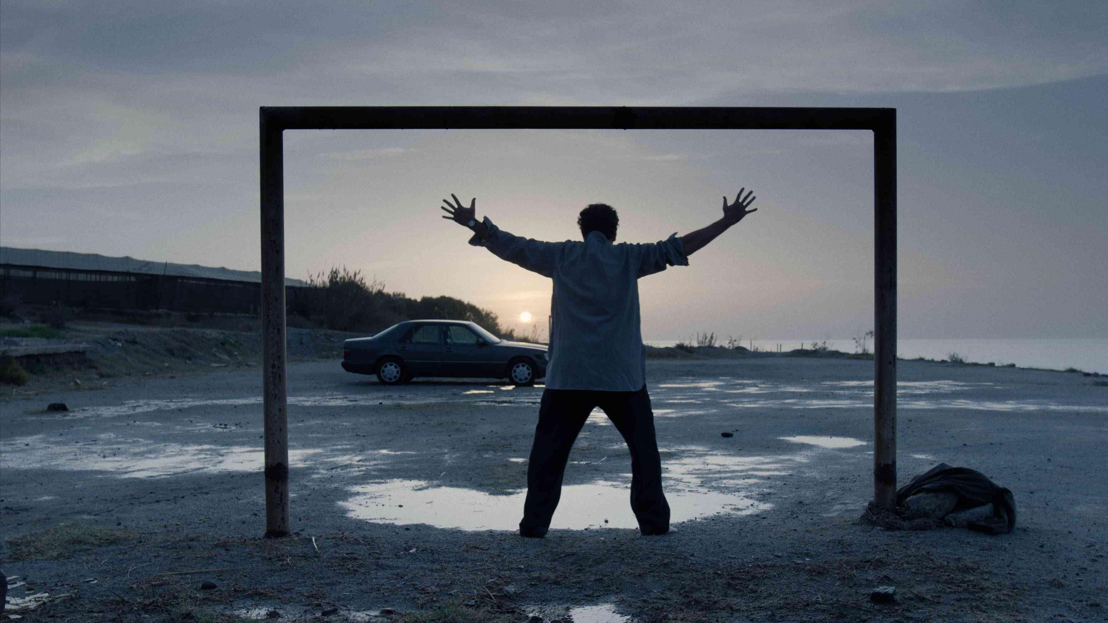 A man with arms outstretch standing in a square frame in an empty parkin lot next to the sea at dawn.