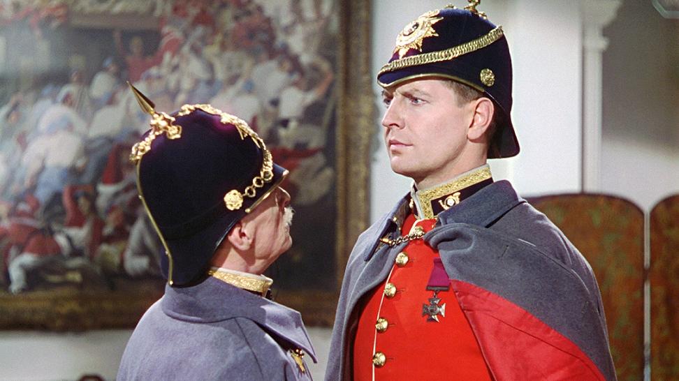 A shorter older solider in a gray uniform addresses a taller young soldier in a red uniform at close range.