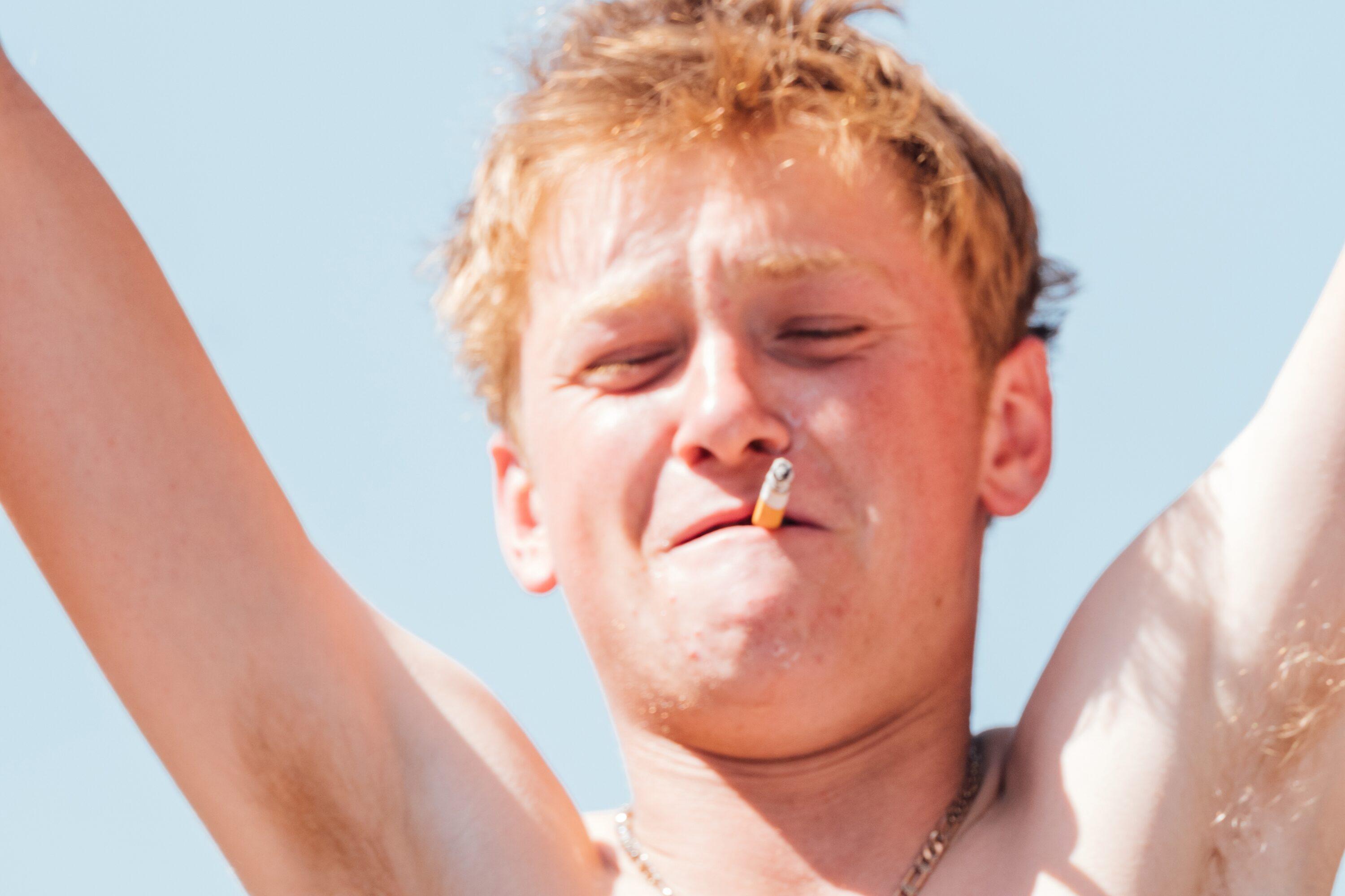 A person with a cigarette in mouth, arms raised, light brown or blonde hair, and a necklace stands against a clear blue sky background.