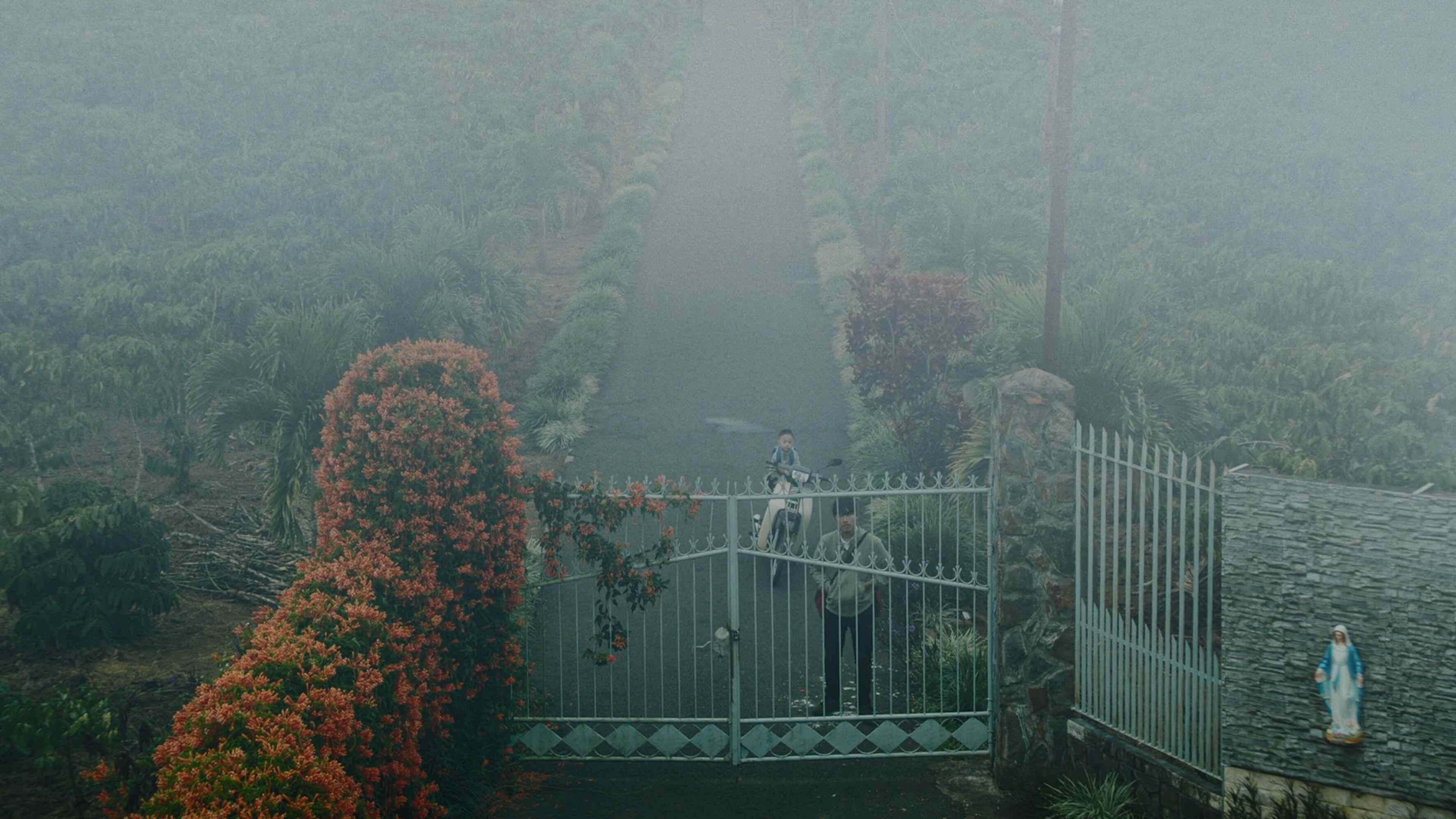 A man stands in front of an iron gate with a child sitting on a motorbike just behind him.