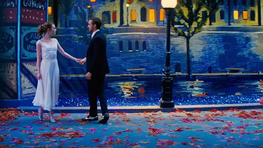 The image shows two people standing on a stage set that resembles a romantic, nighttime cityscape. The person on the left is wearing a white dress, and the person on the right is wearing a black suit. They are holding hands and facing each other. The background features a painted mural of a river with reflections of buildings and trees, illuminated by streetlights. The ground is covered with scattered red and orange leaves, and there are flowers on the right side of the image. The scene is visually striking