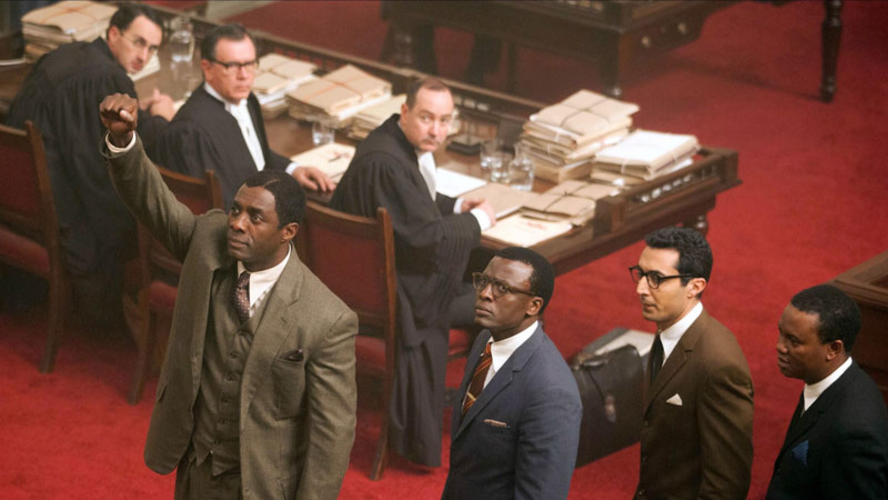 An aerial shot of a man in a suit raising his fist, while others look on in a courtroom with a red rug.