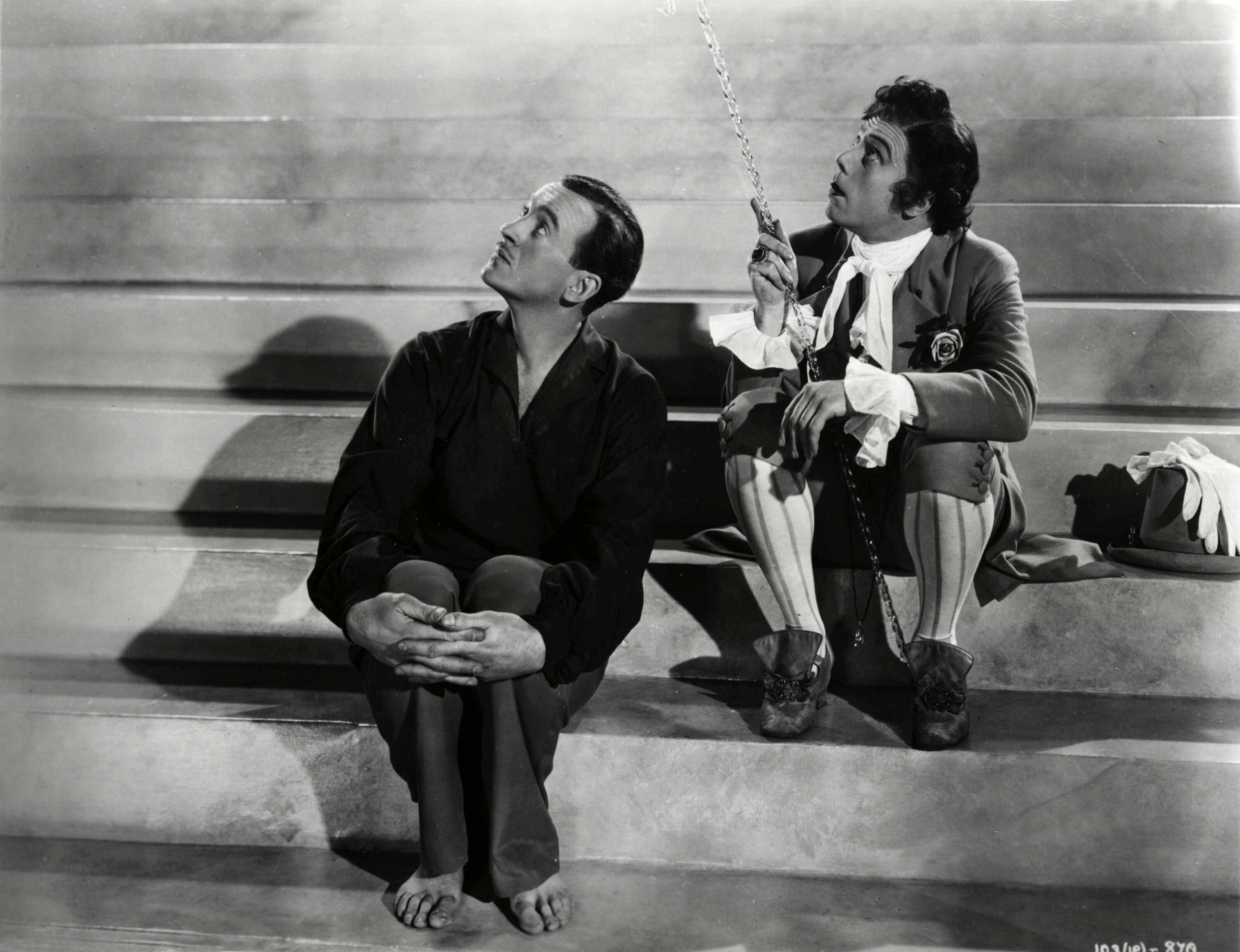 Two individuals sit on a set of steps. The person on the left is barefoot, wearing a dark shirt and pants, and the person on the right wears an elaborate historical costume and holds a cane.