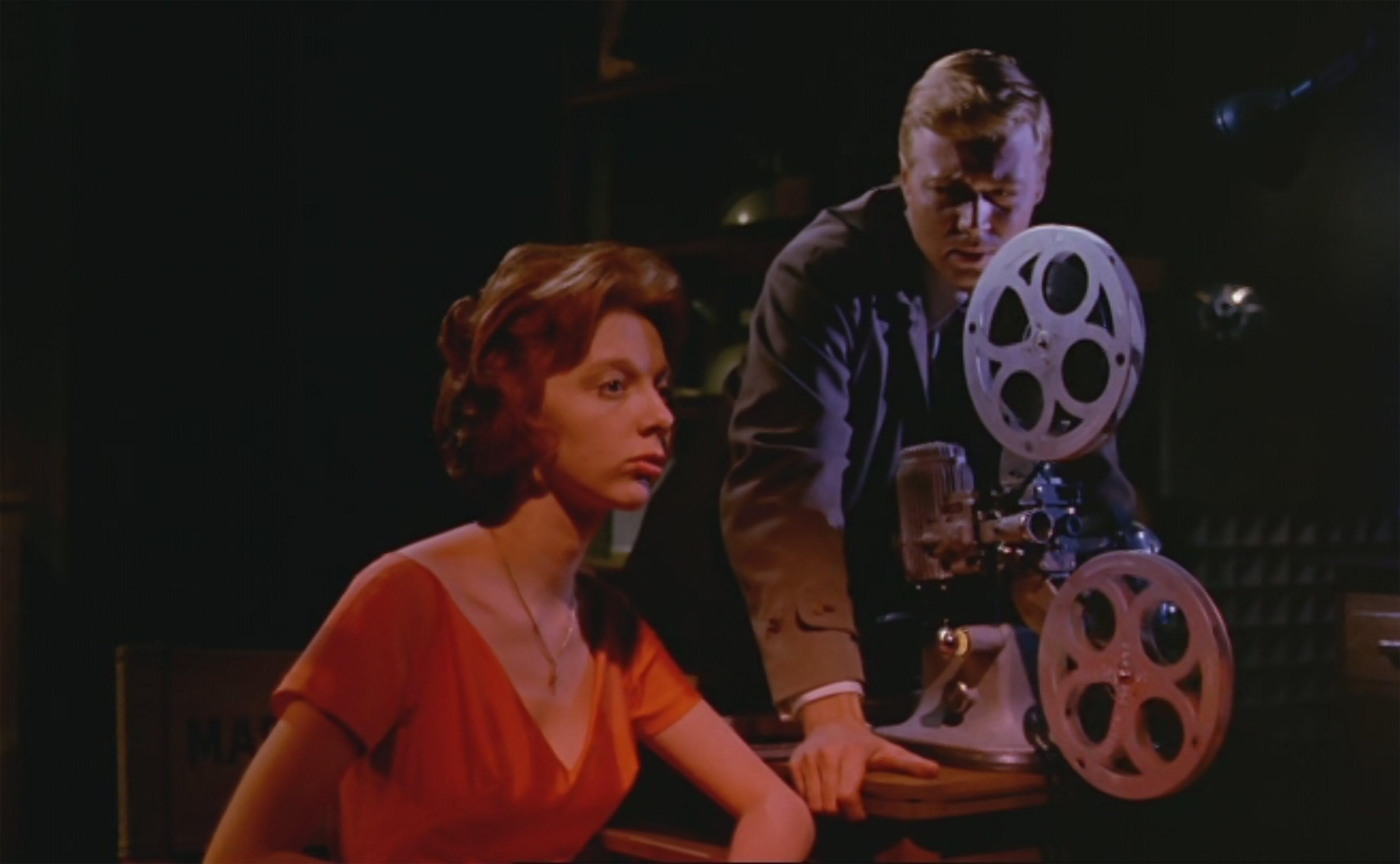 Two individuals are in a dimly lit room; one is seated and wearing a red dress, while the other is standing and operating a vintage film projector.