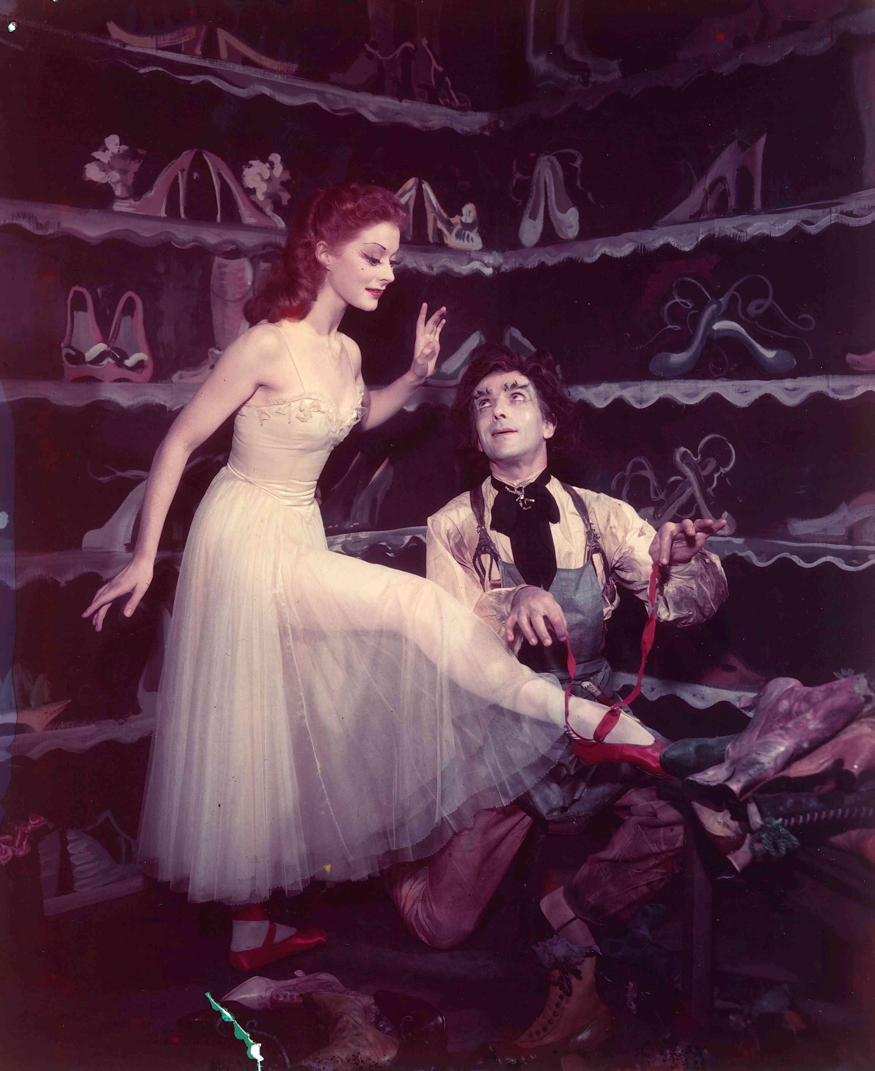 Two individuals in a theatrical setting; one stands in a light-colored dress while the other kneels, assisting with a red shoe, against a backdrop of shoe-filled shelves.