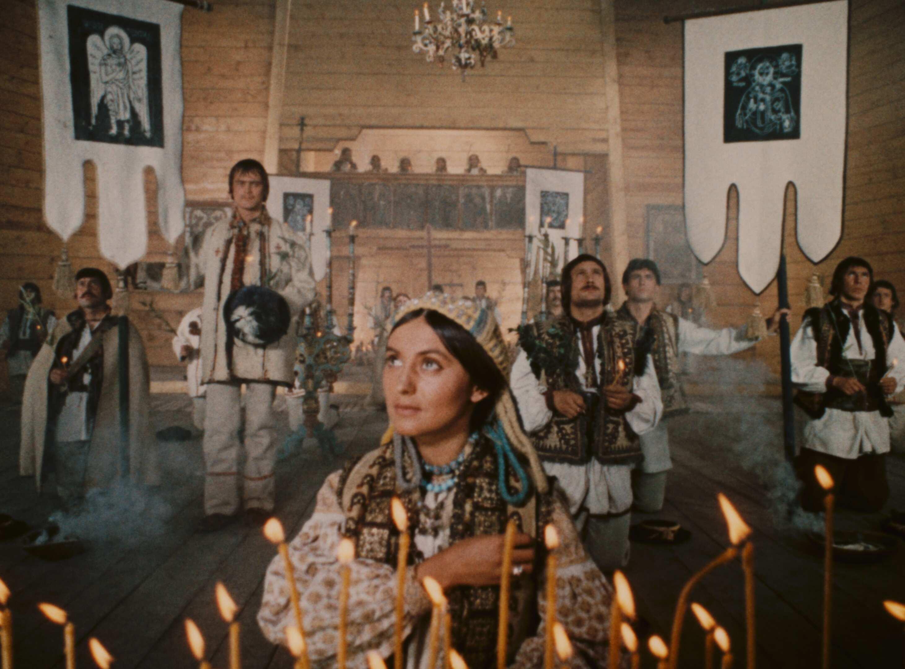 A group of people inside a wooden structure, possibly a church or ceremonial hall, dressed in traditional attire, holding candles amidst smoke, suggesting a religious or cultural ceremony.