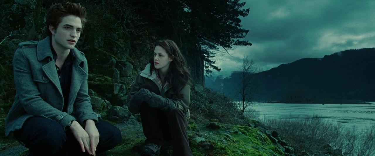 Two people sitting on a moss-covered ground near a body of water with mountains in the background, one in a gray coat and dark pants, and the other in a light-colored jacket and gloves, in a natural, possibly forested area under a cloudy sky.