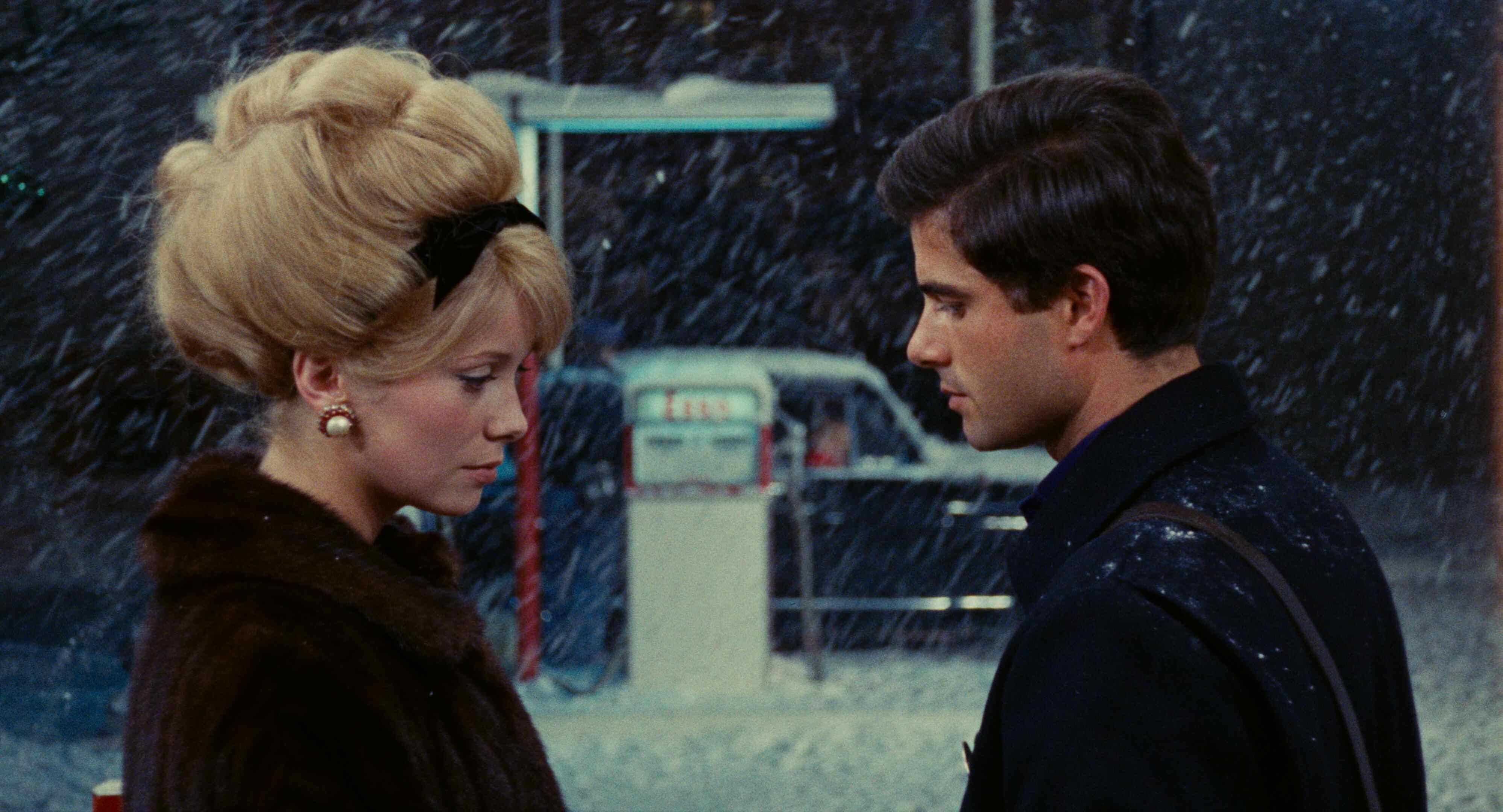 Two individuals standing outside in a snowy environment, near a gas station with falling snow creating a wintry scene.