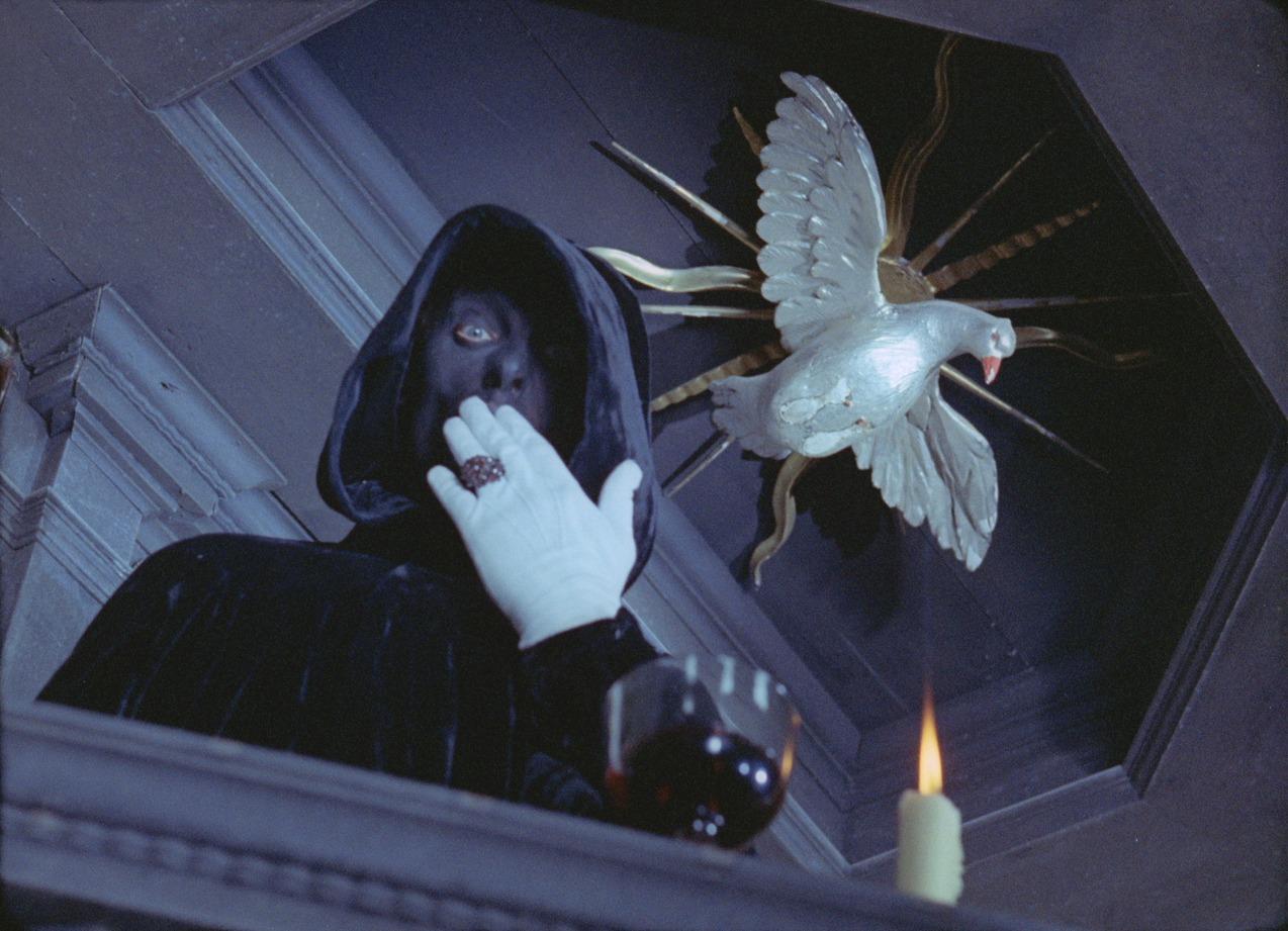 A person dressed in a black hooded cloak, black face mask, white gloves, and large ring, with a lit candle and glass of red liquid in the foreground, and a decorative white bird on a wall in the background.