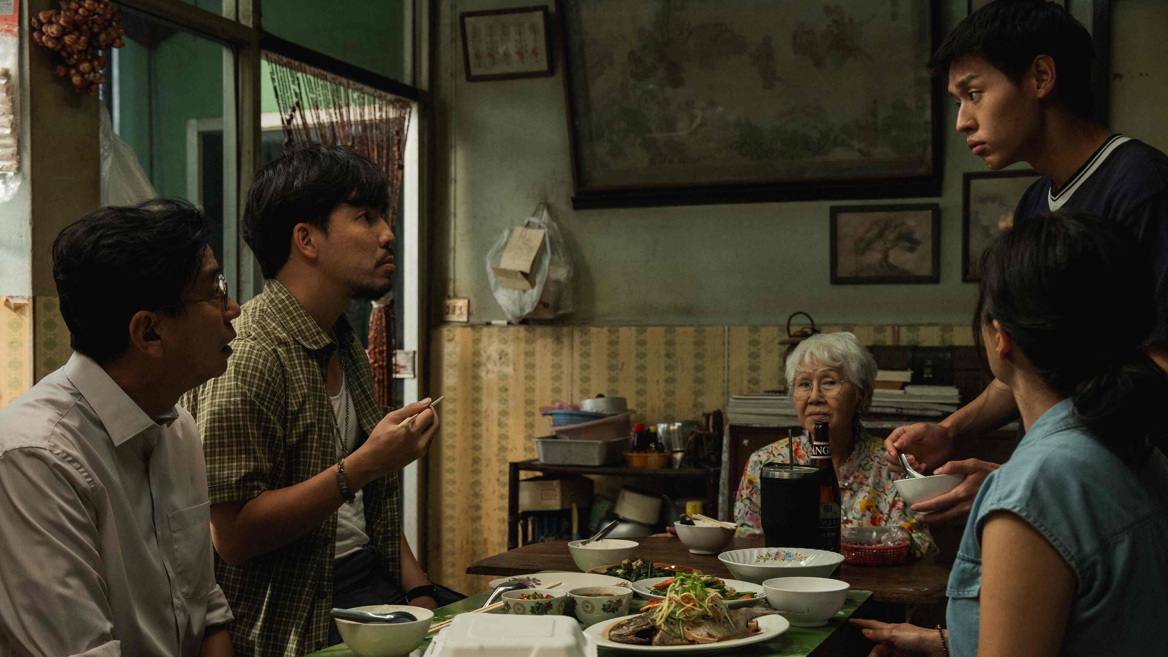 A group of people is gathered around a table, sharing a meal. The table is set with various dishes, including a plate of fish, a bowl of soup, and several smaller bowls containing different foods. The setting appears to be a modest home with a mix of traditional and modern elements, including patterned wallpaper, framed pictures, and a large painting on the wall. A shelf with books and containers is also visible in the background.