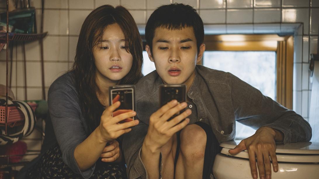 A girl and a boy sit very close together looking at their phones.