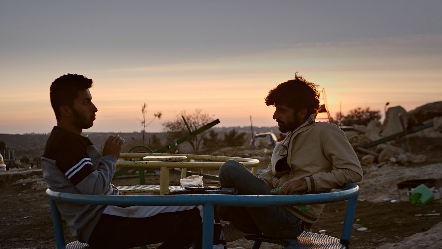 Two men sit facing each other with a sunset and construction debris in the background.