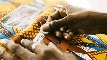 A close up image of hands weaving a colorful yellow, brown, white, and blue geometric fabric.