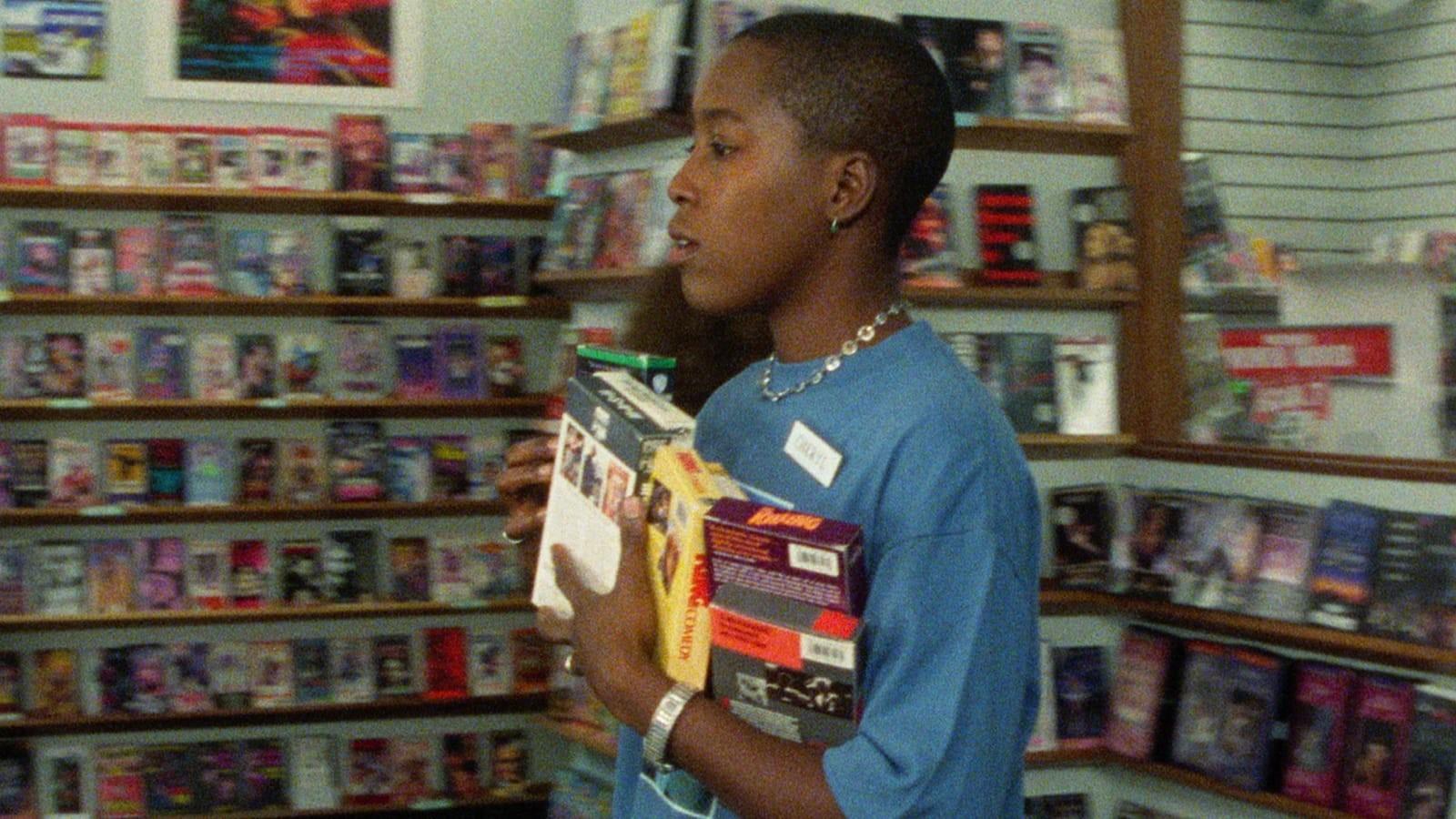A grainy image of woman with a blue shirt carrying an armful of VHS tapes inside a video store.