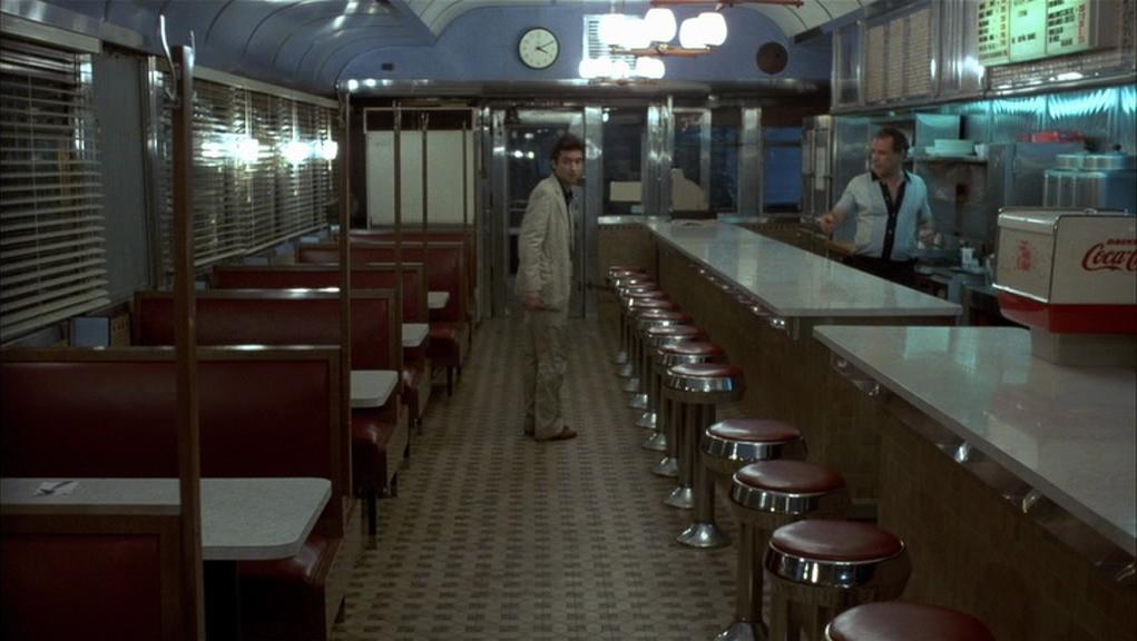 Interior of a classic American diner with red leather booths and red cushioned stools at a counter; a man in a light suit stands facing a uniformed man behind the counter, with a clock and menu boards visible in the background