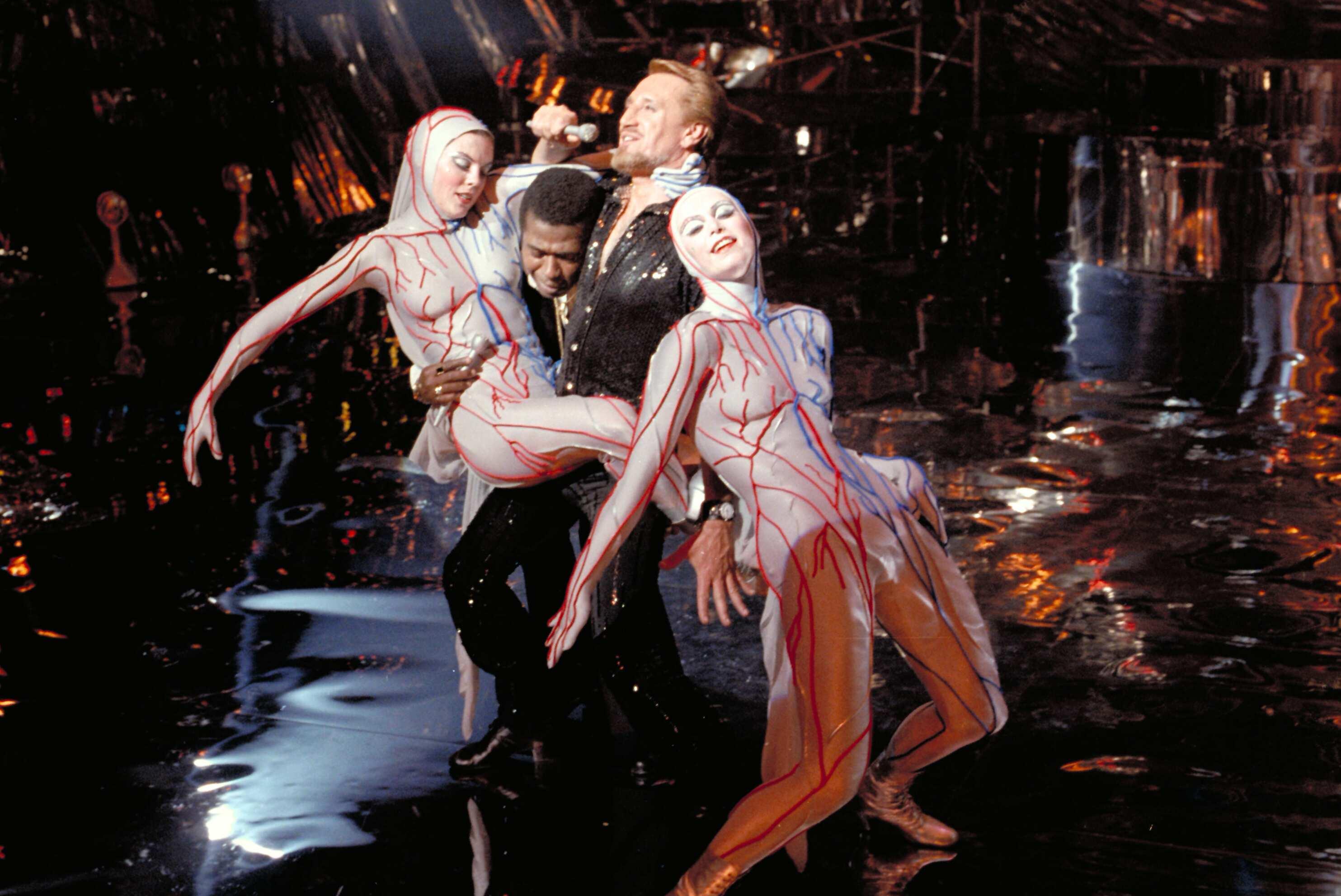 A performer in a black outfit sings into a microphone, flanked by two dancers in red-veined bodysuits striking dramatic poses