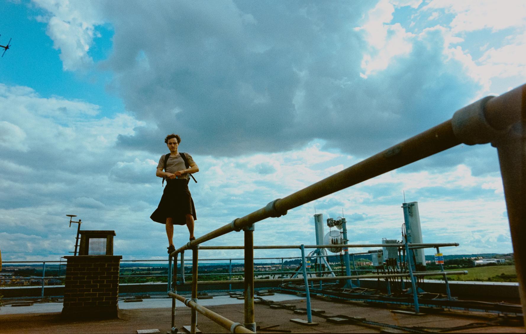 Person standing on a metal railing on a rooftop, with an industrial area and cloudy sky in the background