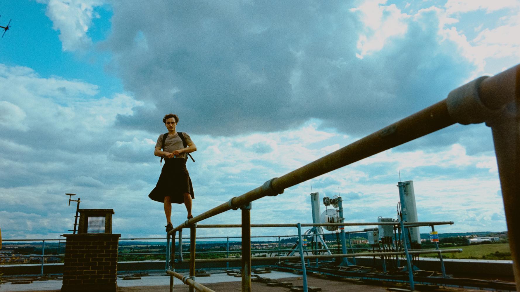Person standing on a metal railing on a rooftop, with an industrial area and cloudy sky in the background
