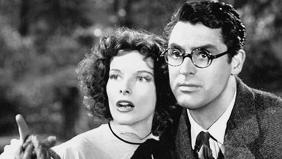 Two people stand close together outdoors; one wears a ruffled dress and holds a coiled rope, the other wears a suit and tie