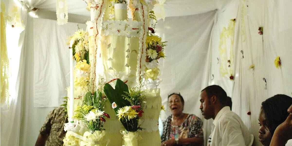 A floral and candle-decorated structure surrounded by seated individuals, set against white drapes and more floral arrangements