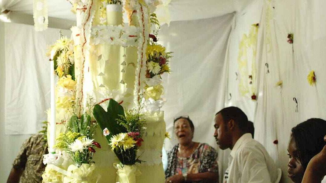 A floral and candle-decorated structure surrounded by seated individuals, set against white drapes and more floral arrangements