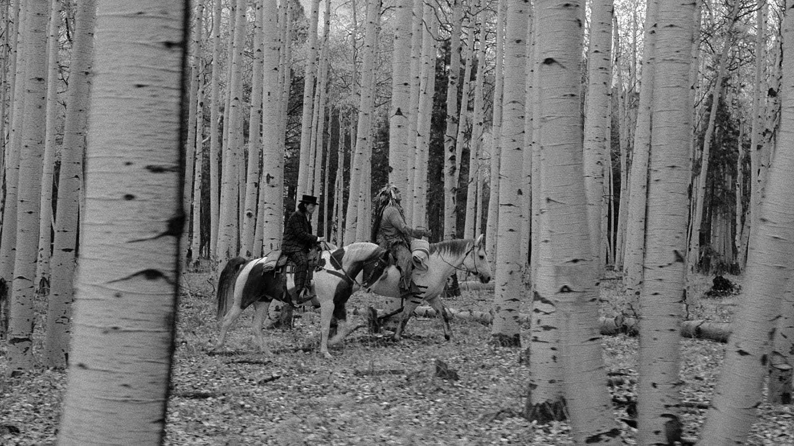Black and white image of two individuals riding horses through a forest of tall, slender trees. The forest floor is covered with fallen leaves and no visible trail