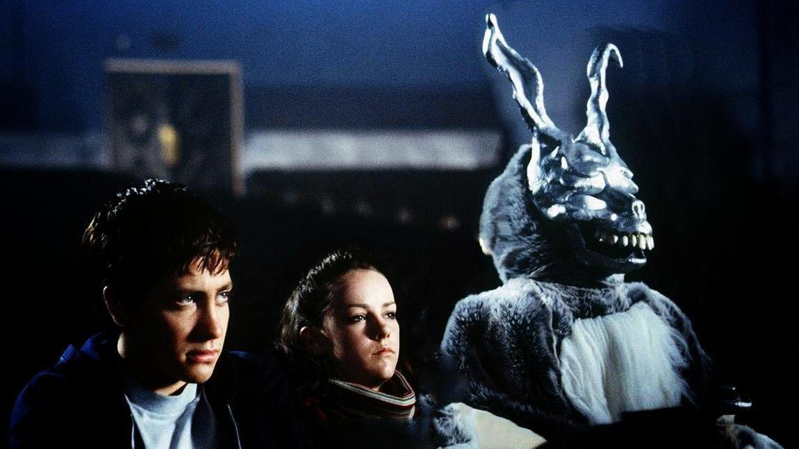 Three individuals sit in a dimly lit setting; one wears a large rabbit costume with sharp teeth and prominent ears