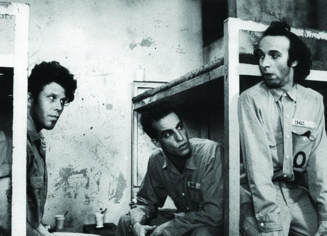 Three individuals in a prison room with bunk beds and graffiti on the wall; one wears a uniform labeled '19421'. Two cups sit on a nearby surface