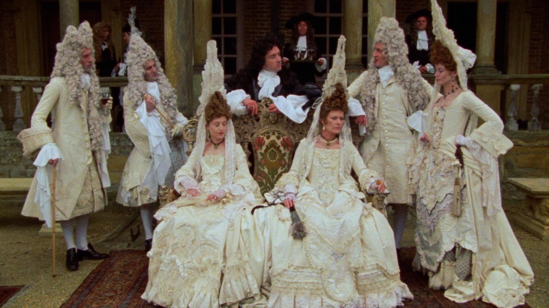 Group of people in ornate 17th or 18th-century costumes with wigs, posed in front of a columned building