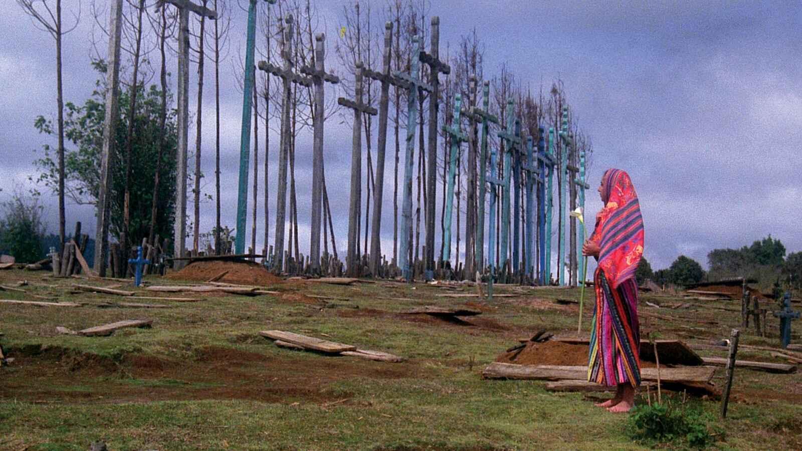 A person wrapped in a colorful blanket stands in front of a field of tall wooden crosses, holding an object possibly a candle or flower. The sky is overcast and the ground is uneven