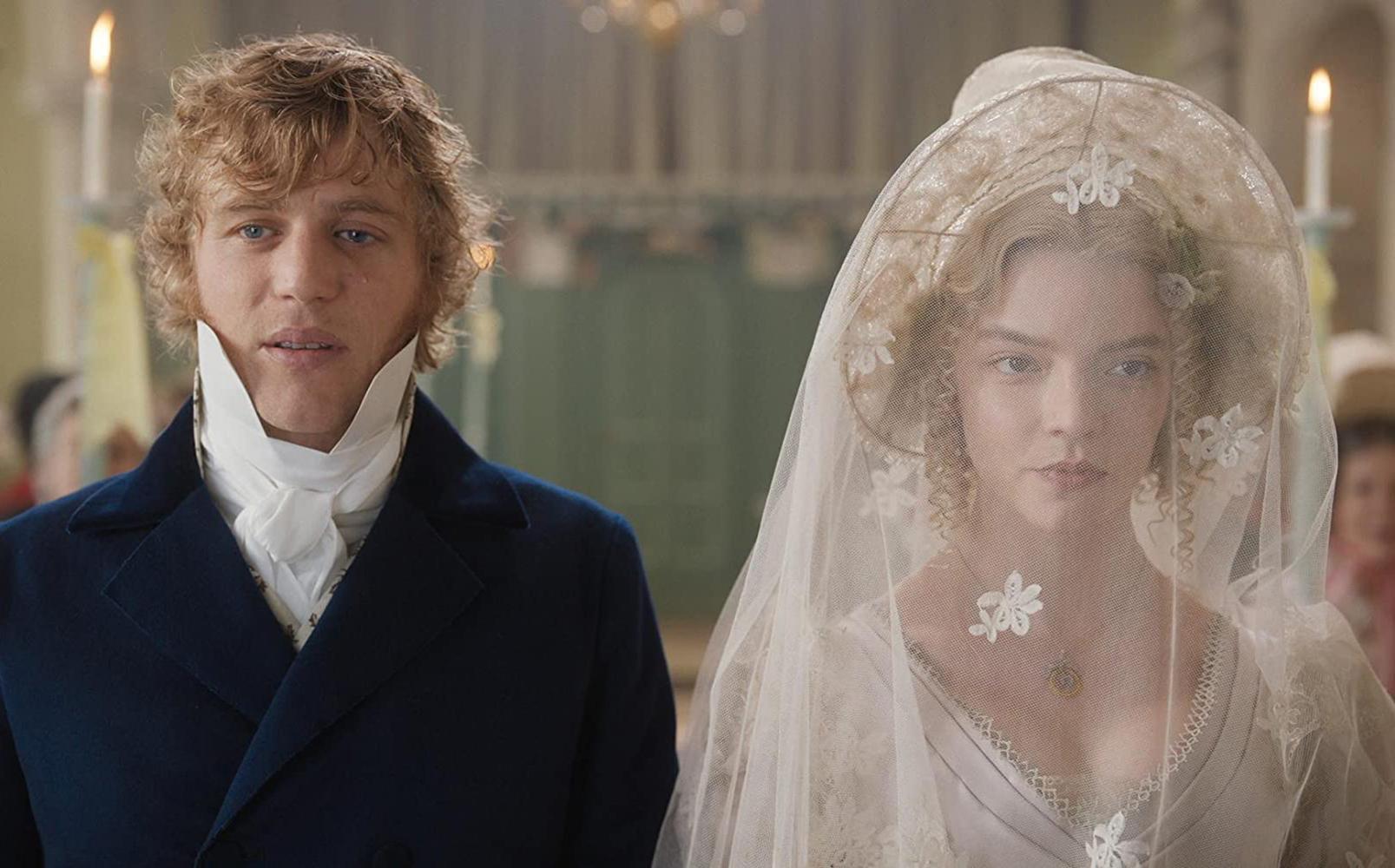 Two people in historical attire stand side by side indoors; one wears a dark blue coat with a white cravat, the other a white wedding dress with a lace veil