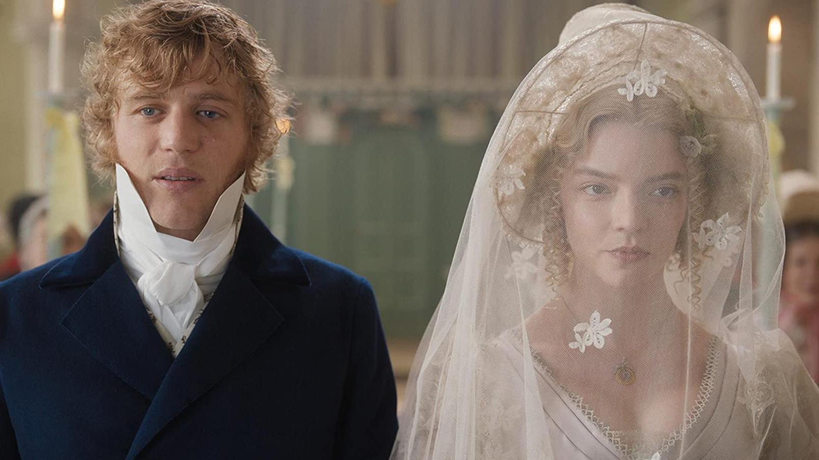 Two people in historical attire stand side by side indoors; one wears a dark blue coat with a white cravat, the other a white wedding dress with a lace veil