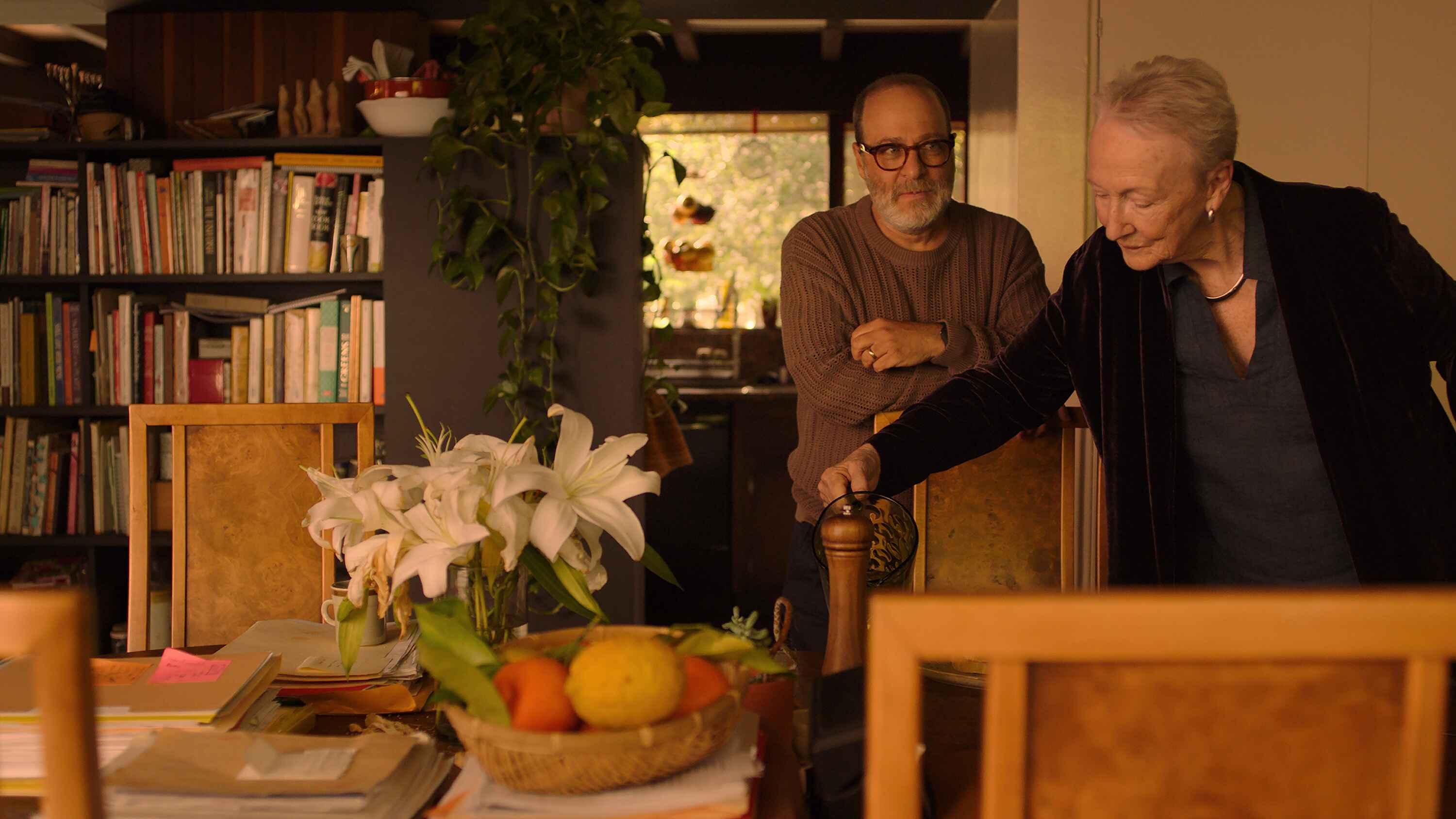 Two people stand near a table holding a basket of fruits, white lilies in a vase, and papers. A bookshelf filled with books and plants is visible in the background