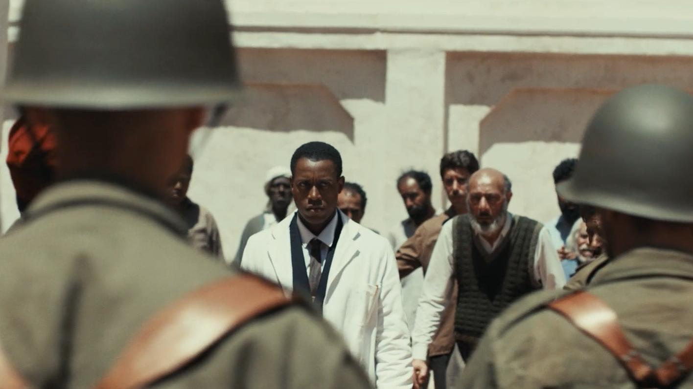 Group of people in an outdoor setting; two in the foreground wear military helmets and uniforms, while a person in a white lab coat stands among civilians in the background