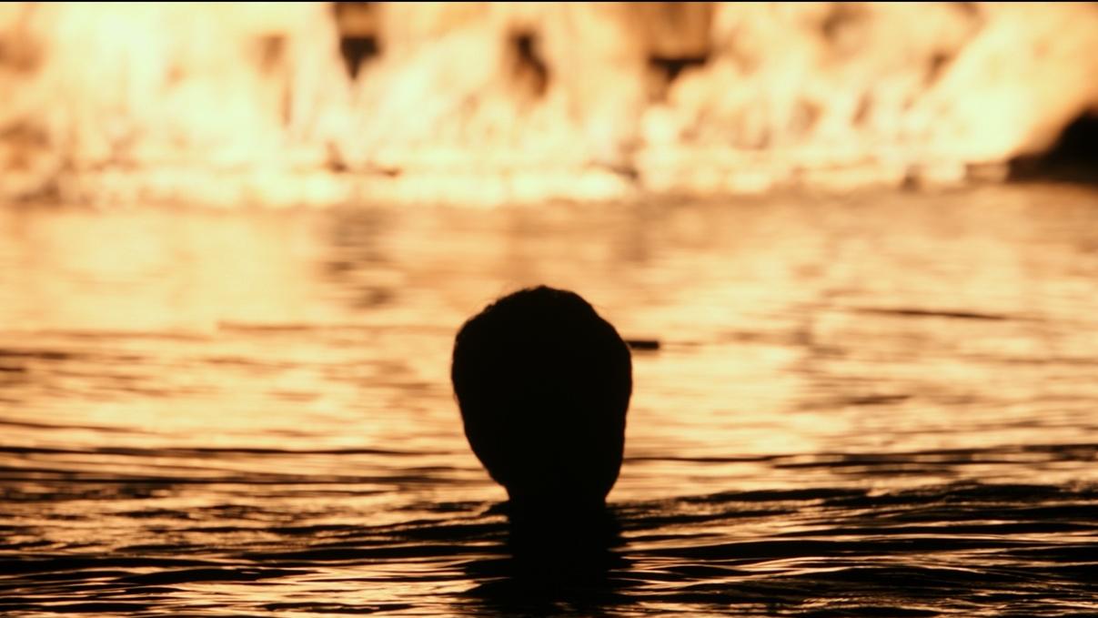 A shadowy image of a boy in the water looking at a boat on fire.