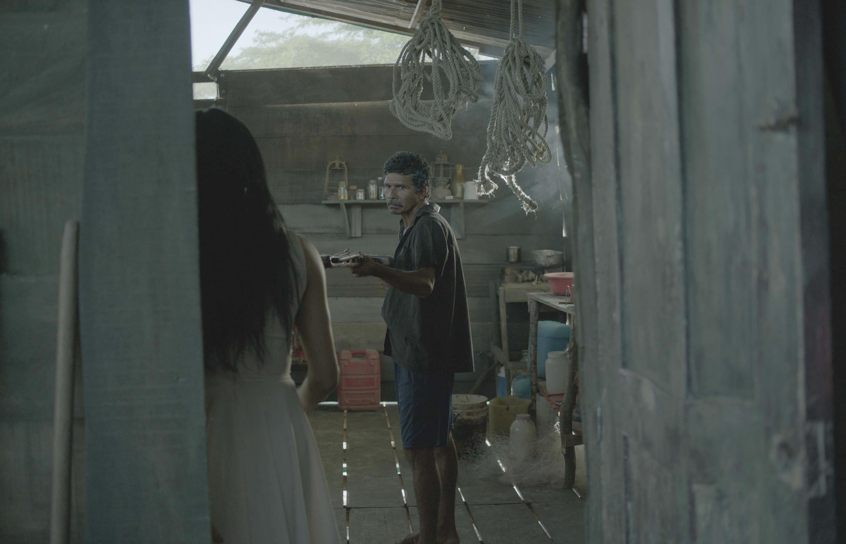 Inside a dimly lit rustic room with wooden walls and floor, one person stands holding an object while another is partially visible in the foreground; ropes hang from the ceiling and shelves line the back wall
