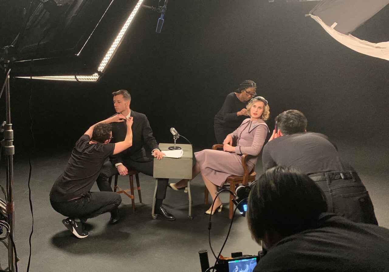 Behind-the-scenes setup of a photoshoot or video recording with two seated individuals and a microphone between them. Crew members adjust lighting and equipment in a dark setting