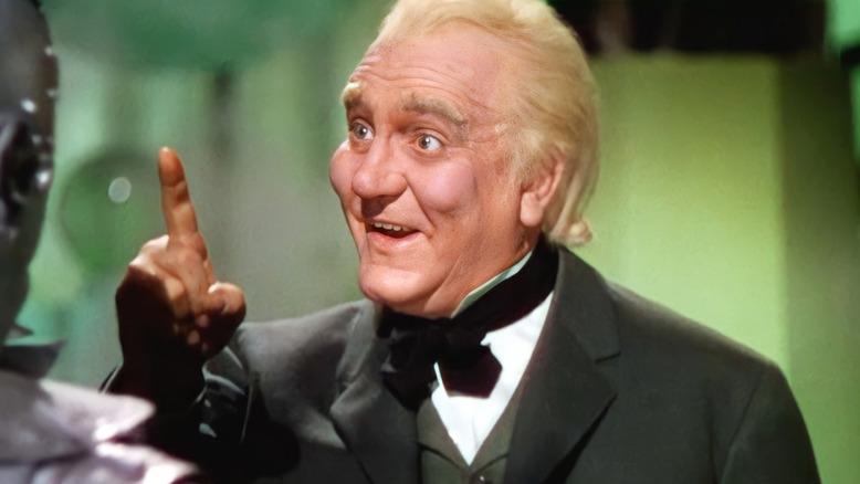 A cheerful man with blonde hair and wearing a dark green suit with a bow tie gestures with a pointed finger.