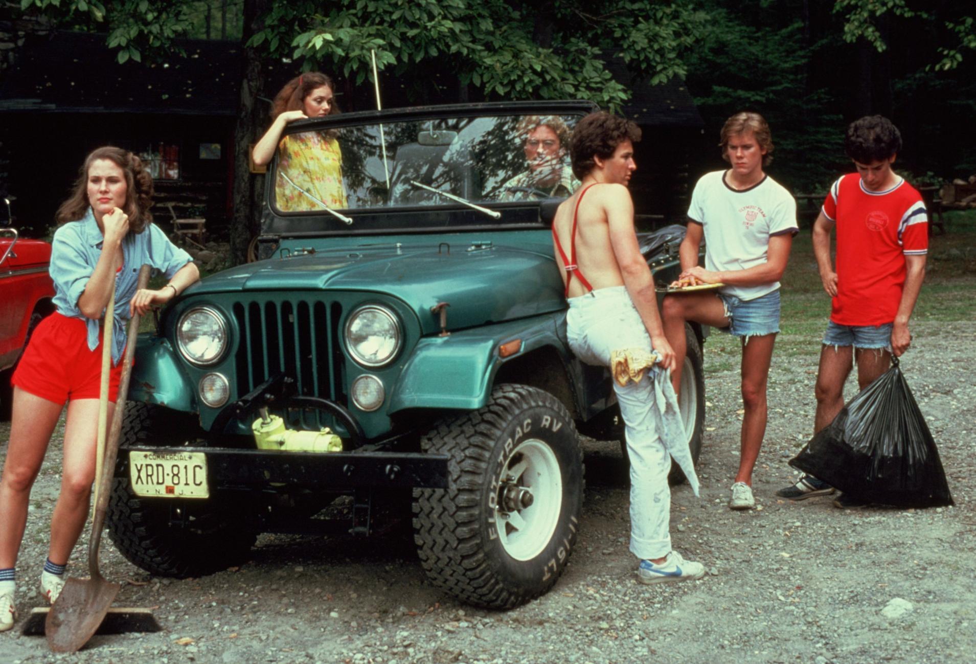 A group of people gather around a green Jeep; one person leans on the front holding a shovel, another sits on the hood