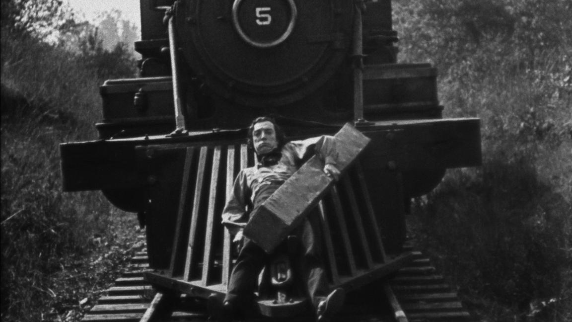 A black-and-white photo of a man clinging to the front of a steam engine train and holding a large block of wood.