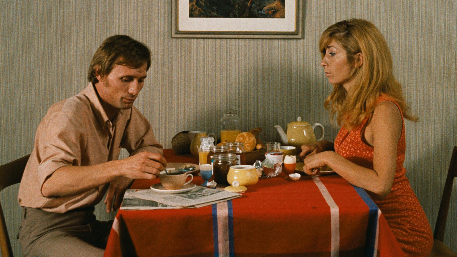Two people sit at a breakfast table with a red, white, and blue striped tablecloth, surrounded by cups, a teapot, jam, juice, bread, and a newspaper; striped wallpaper and a framed picture are in the background