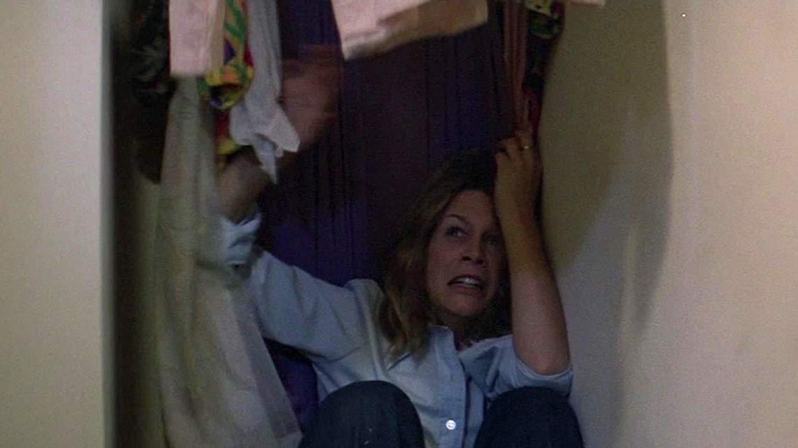 A frightened person sits hiding on the floor of a closet surrounded by hanging clothes