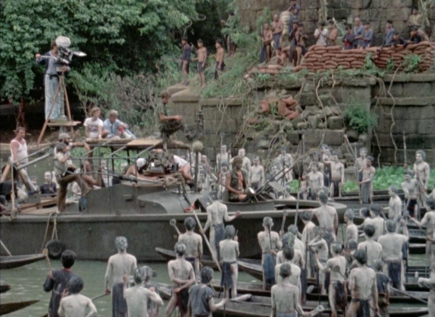 A movie set scene with a boat carrying crew and equipment in the water, surrounded by people in small boats or standing in water, many covered in white paint; an ancient stone structure with vegetation is in the background