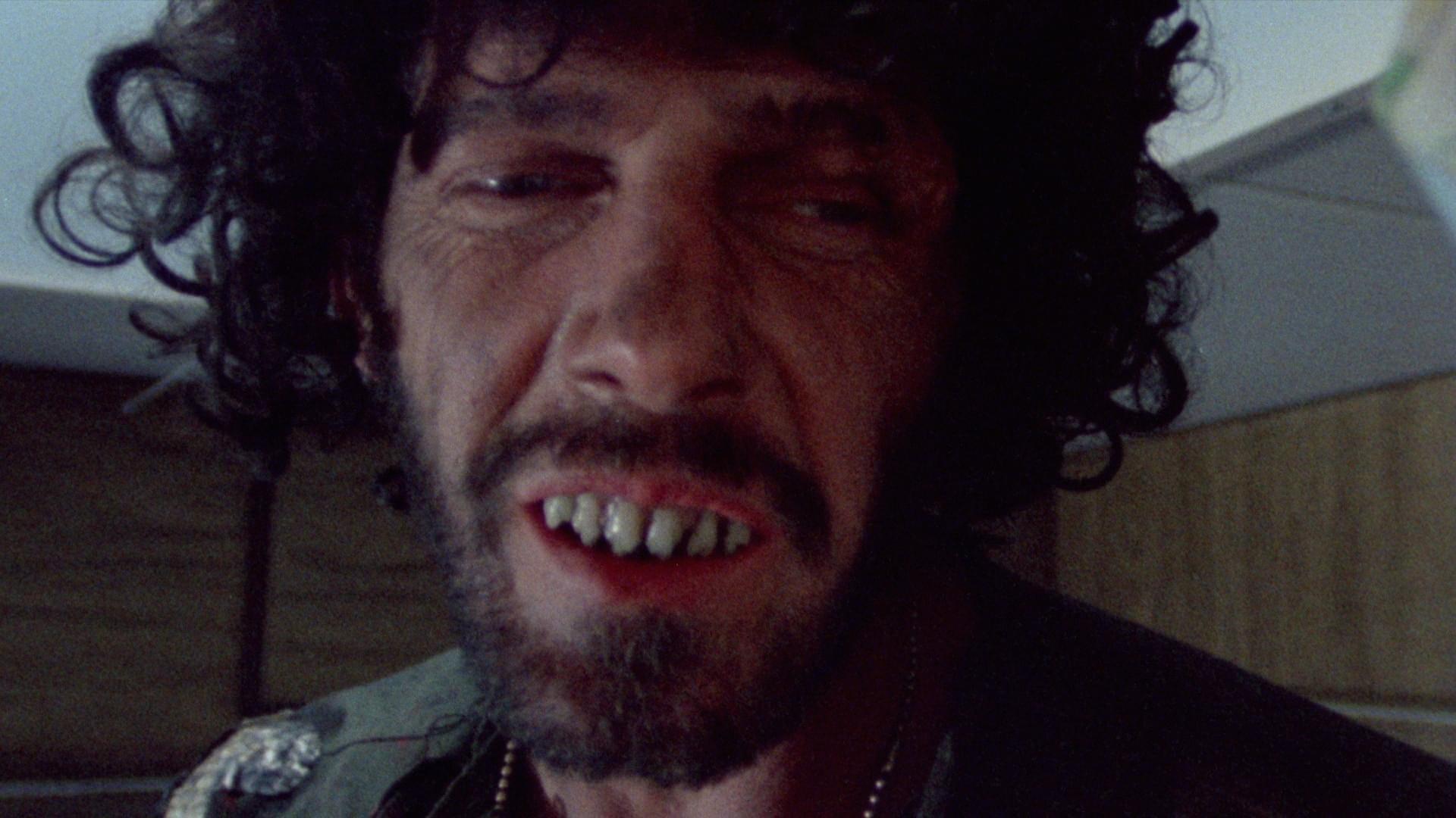 Close up image of a person's face with black curly hair, facial hair, and exposed teeth.
