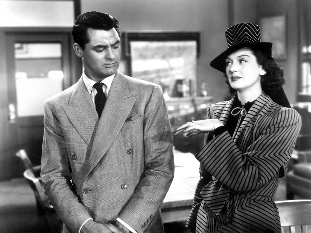 Black and white photo of two individuals in an office setting; one wears a double-breasted suit with a tie, the other a striped outfit with a hat