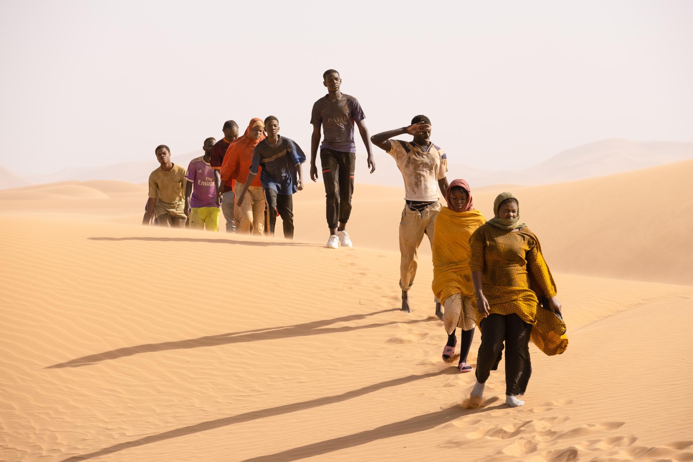 Ten individuals walk in a line across a desert landscape with rolling sand dunes under a clear sky